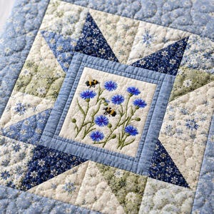 以下が含まれることがあります： 青いヤグルマギクと2匹の蜂が刺繍された、中央のデザインが施されたキルトの正方形。 正方形は、青と白の色合いの星模様と花柄で囲まれています。