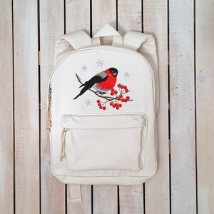 Pu&ograve; includere: Zaino in tela bianca con un ricamo di un uccello dal petto rosso appollaiato su un ramo con bacche rosse e fiocchi di neve.