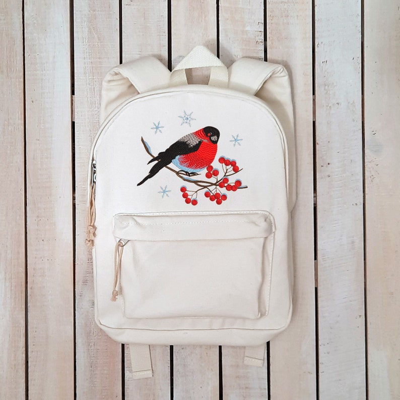 Pu&ograve; includere: Zaino in tela bianca con un ricamo di un uccello dal petto rosso appollaiato su un ramo con bacche rosse e fiocchi di neve.