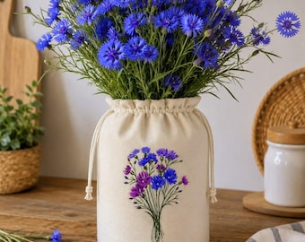 May include: A bouquet of vibrant blue cornflowers in a cream-colored drawstring vase. The vase features an embroidered floral design in shades of blue and purple. The arrangement sits on a wooden surface, with a cutting board and small plant in the background.