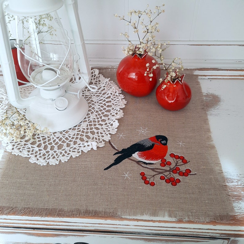Pu&ograve; includere: Un runner da tavolo in lino marrone con un pizzo all'uncinetto bianco, due melograni in ceramica rossa e un'immagine ricamata di un pettirosso appollaiato su un ramo con bacche rosse.