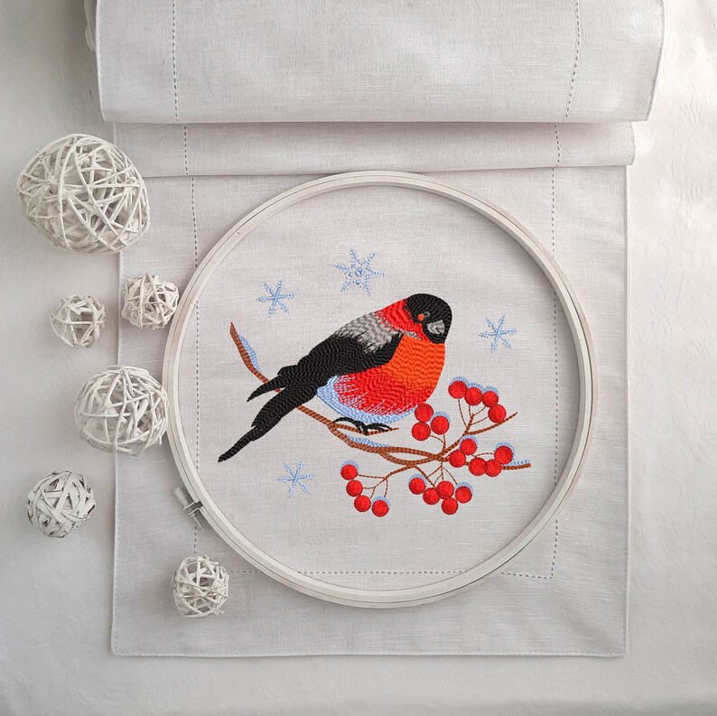 Pu&ograve; includere: Arte di ricamo in cerchio con un pettirosso dal petto rosso appollaiato su un ramo con bacche rosse. L'uccello &egrave; circondato da fiocchi di neve su uno sfondo di lino bianco.