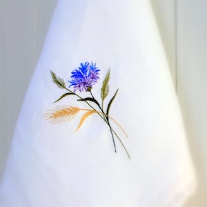 Könnte beinhalten: Ein weißes Geschirrtuch mit einer gestickten blauen und violetten Blume und grünen Stielen. Die Blume ist von einigen kleinen goldenen Weizenähren umgeben.