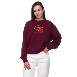 Pu&ograve; includere: Una felpa color bordeaux con un ricamo a forma di uccello rosso su un ramo con bacche rosse e fiocchi di neve.