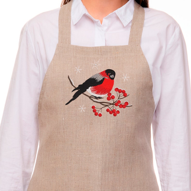 Pu&ograve; includere: Un grembiule in lino beige con un ricamo a forma di pettirosso appollaiato su un ramo con bacche rosse. Il disegno &egrave; circondato da fiocchi di neve.