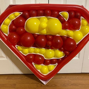 May include: A red and yellow balloon sculpture in the shape of the Superman logo. The sculpture is made of many individual balloons, and is displayed on a white background.