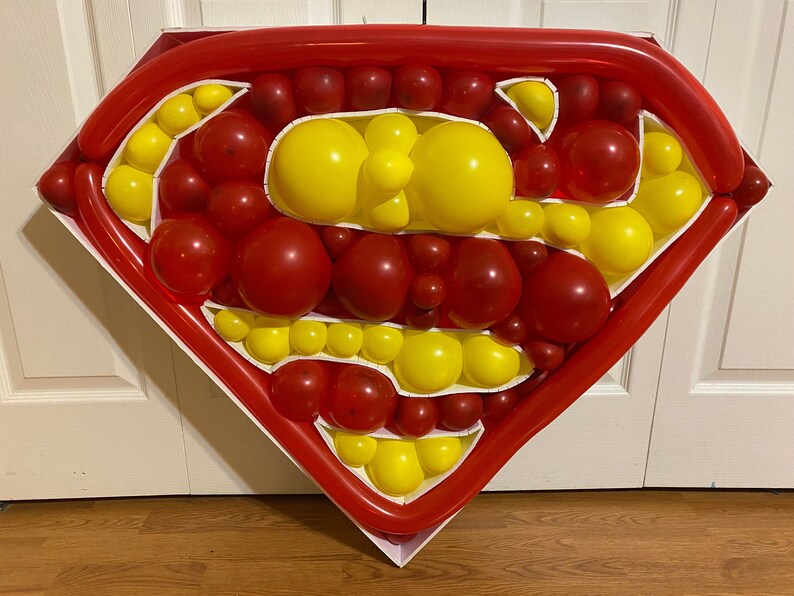 May include: A red and yellow balloon sculpture in the shape of the Superman logo. The sculpture is made of many individual balloons, and is displayed on a white background.
