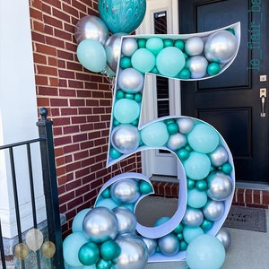May include: A large white number five balloon arch decorated with silver and teal balloons. The balloon arch is standing in front of a brick wall and a black door.