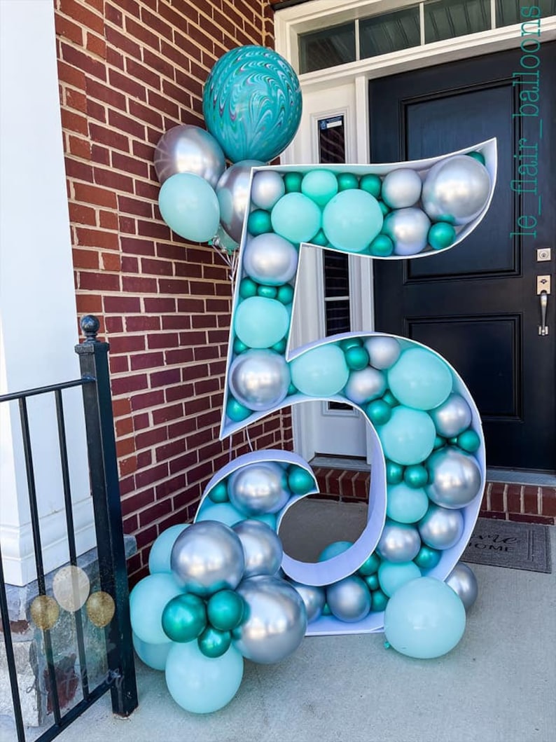 May include: A large white number five balloon arch decorated with silver and teal balloons. The balloon arch is standing in front of a brick wall and a black door.