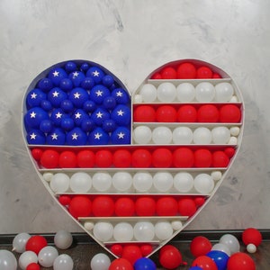 May include: A heart-shaped frame filled with red, white and blue balloons, forming the American flag. Balloons are scattered on the floor.