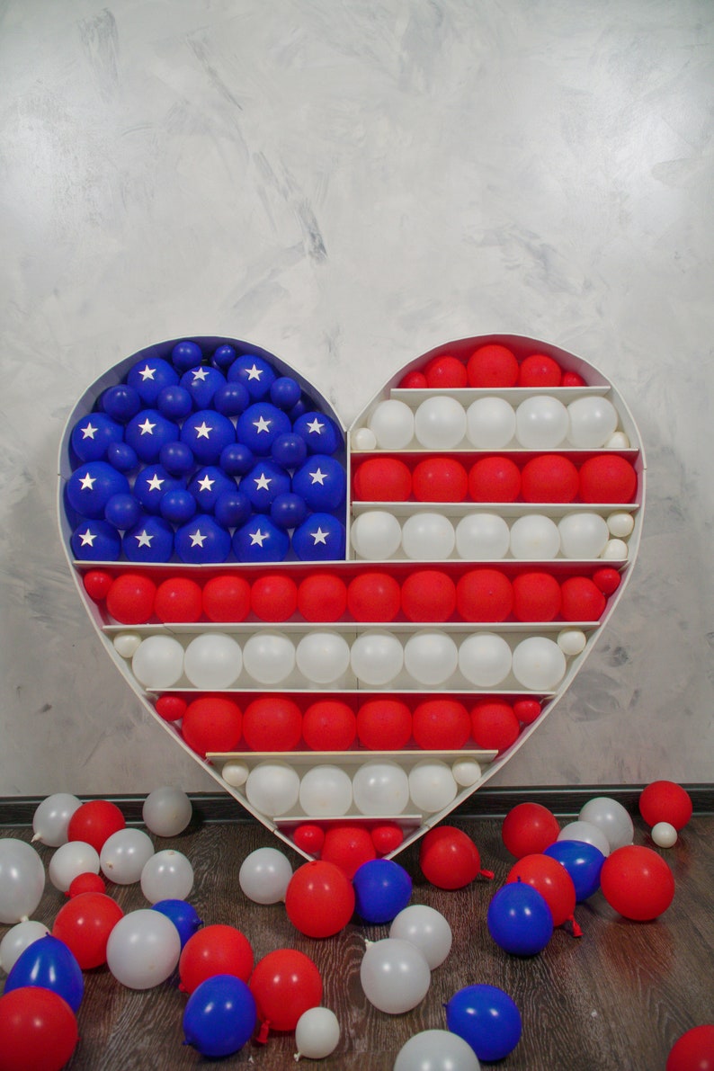 May include: A heart-shaped frame filled with red, white and blue balloons, forming the American flag. Balloons are scattered on the floor.