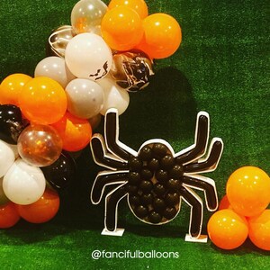 May include: A black balloon spider decoration with a white outline and black balloons for the body. The spider is surrounded by orange and gray balloons.