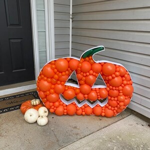 May include: A large orange pumpkin made of balloons with a jack-o'-lantern face. The pumpkin is sitting on a concrete porch with a welcome mat in front of a door.