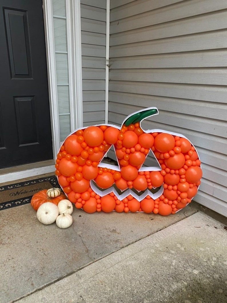 May include: A large orange pumpkin made of balloons with a jack-o'-lantern face. The pumpkin is sitting on a concrete porch with a welcome mat in front of a door.