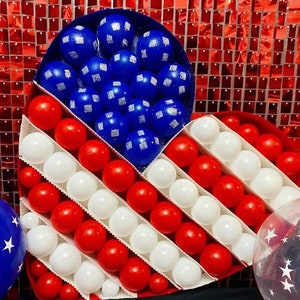May include: A heart-shaped balloon arrangement in red, white and blue, resembling the American flag. The balloons are arranged in a grid pattern, with red and white balloons alternating in rows. The heart is surrounded by a red, white and blue sequin backdrop.