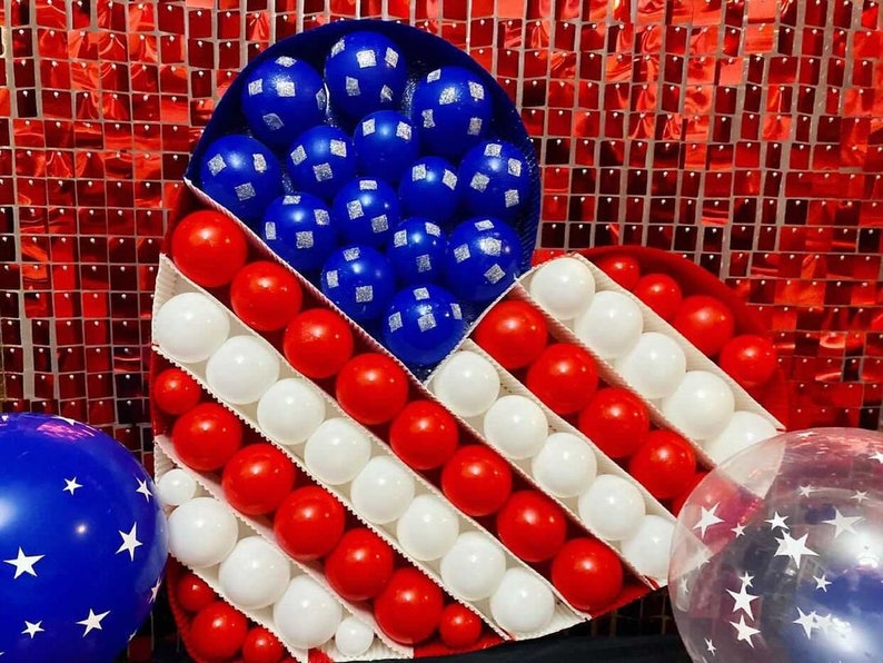 May include: A heart-shaped balloon arrangement in red, white and blue, resembling the American flag. The balloons are arranged in a grid pattern, with red and white balloons alternating in rows. The heart is surrounded by a red, white and blue sequin backdrop.