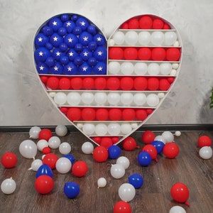 May include: A white heart-shaped frame filled with red, white and blue balloons, arranged in the pattern of the American flag. Some balloons have fallen to the floor.