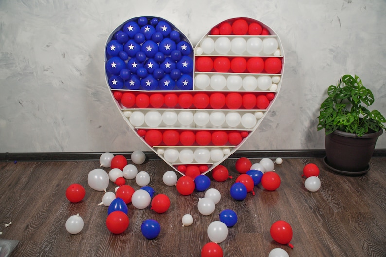 May include: A white heart-shaped frame filled with red, white and blue balloons, arranged in the pattern of the American flag. Some balloons have fallen to the floor.