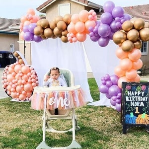 May include: A baby girl sits in a high chair with a peach and gold balloon arch behind her. The high chair has a banner that says "one" on it. There is a chalkboard sign that says "Happy Birthday Nora 1" with a pumpkin design.