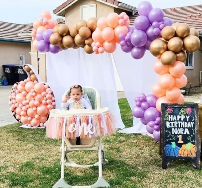 May include: A baby girl sits in a high chair with a peach and gold balloon arch behind her. The high chair has a banner that says "one" on it. There is a chalkboard sign that says "Happy Birthday Nora 1" with a pumpkin design.