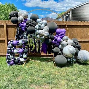 May include: A large number six balloon arch made of black, white and purple balloons. The balloons are arranged in a circular shape and are decorated with streamers. The arch is set up in front of a wooden fence.