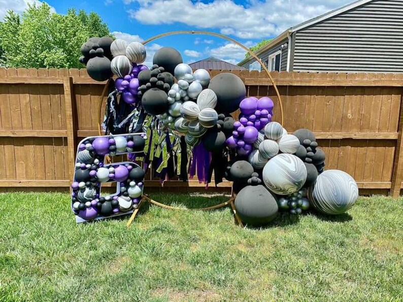 May include: A large number six balloon arch made of black, white and purple balloons. The balloons are arranged in a circular shape and are decorated with streamers. The arch is set up in front of a wooden fence.