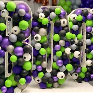 May include: A large BOO sign made of balloons in green, purple, black, white, and silver. The balloons are arranged to form the letters BOO. Some of the balloons have googly eyes.