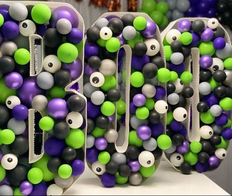 May include: A large BOO sign made of balloons in green, purple, black, white, and silver. The balloons are arranged to form the letters BOO. Some of the balloons have googly eyes.