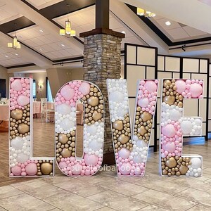 May include: White, pink, and gold balloon letters spelling "LOVE" in a large, decorative font. The letters are filled with balloons and are standing on a tiled floor.