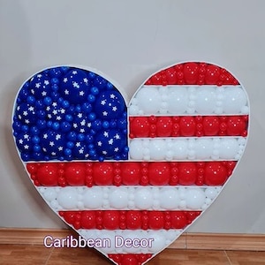 May include: A heart-shaped balloon arrangement in the shape of the American flag. The arrangement is made of red, white, and blue balloons. The stars are white on a blue background. The stripes are red and white. The arrangement is on a wooden floor. Caribbean Decor is written in pink at the bottom of the image.
