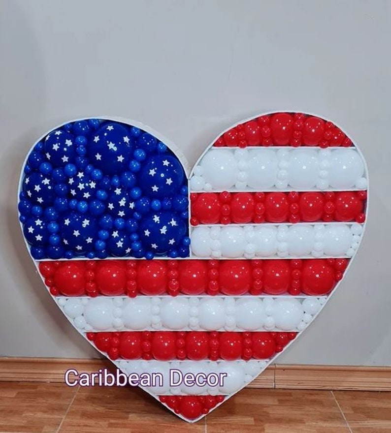 May include: A heart-shaped balloon arrangement in the shape of the American flag. The arrangement is made of red, white, and blue balloons. The stars are white on a blue background. The stripes are red and white. The arrangement is on a wooden floor. Caribbean Decor is written in pink at the bottom of the image.