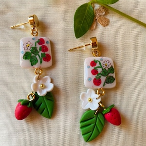 Può includere: Orecchini dorati con un motivo a fragole sulla parte superiore quadrata, un fiore bianco, una foglia verde e un ciondolo a fragola rossa. Chiusura a perno.