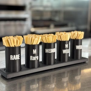 Könnte beinhalten: Ein schwarzes Holztablett hält fünf schwarze zylindrische Behälter mit Holz-Steakmarkern. Jeder Behälter ist mit einem anderen Garstufen-Namen beschriftet: RARE, M Rare, MEDIUM, M WELL und WELL. Die Marker sind hellgelb.