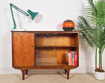 May include: A mid-century modern wooden cabinet with a door and glass-fronted shelves. A green desk lamp and a decorative ball sit on top. A potted palm tree is to the right.