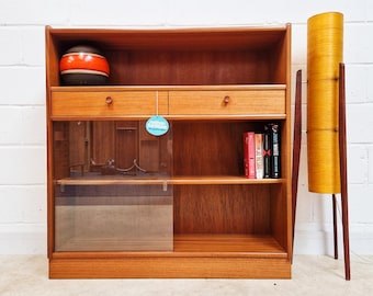 May include: A teak wood bookcase featuring an open top shelf, two drawers with circular wooden pulls, and a lower section with a sliding glass door. A round blue tag with the text Nathan Furniture is attached. A tall cylindrical floor lamp stands nearby.