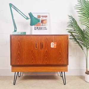 May include: A mid-century wooden cabinet with two doors and hairpin legs. A green desk lamp and a framed "G Plan" poster sit on top. A tall green plant in a white pot is to the right. The cabinet is approximately 80cm wide.