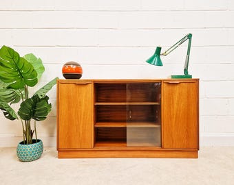 Könnte beinhalten: Ein Mid-Century-Modern-Holzschrank mit zwei Türen und einer Glasschiebetür in der Mitte. Eine grüne Schreibtischlampe und eine dekorative orangefarbene und braune Kugel stehen obenauf. Links eine große Topfpflanze mit grünen Blättern.