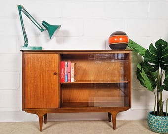 May include: A mid-century modern wooden cabinet with a glass-fronted shelf and a solid door. A green desk lamp and a decorative ball sit atop the cabinet. A potted plant with large green leaves is to the right.