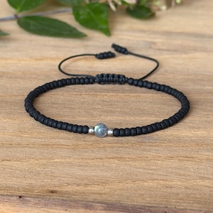 May include: A black beaded bracelet featuring a single central grey speckled stone bead flanked by two small silver-toned metallic beads. The bracelet is constructed with a black cord and includes an adjustable sliding knot closure mechanism.