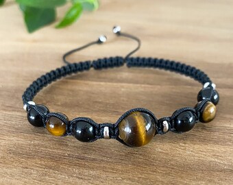 May include: Black macramé bracelet with a large tiger's eye bead and black onyx beads. The adjustable bracelet has silver-coloured accents. The bracelet is displayed on a wooden surface. The bracelet is a piece of jewellery.