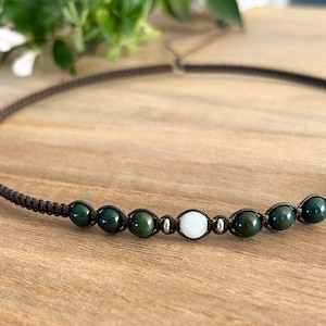 May include: A brown cord necklace with green and white beads. The necklace is made with a simple knot design.