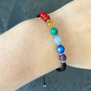 May include: A black cord bracelet with seven colorful round beads. The beads are red, orange, yellow, green, blue, indigo, and purple.