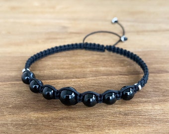 May include: A black cord bracelet featuring a series of smooth, round, polished black beads arranged in a row. The bracelet includes small silver-toned metal accents between the beads and utilises a sliding knot closure with two dangling ends.
