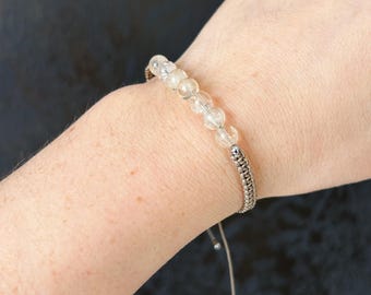 May include: Clear beaded bracelet with a silver chain accent and adjustable cord closure. The bracelet features multiple clear, round beads and a woven silver chain section. The cord is a neutral colour.