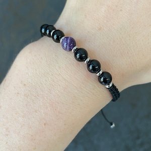 May include: A black beaded bracelet with a purple gemstone bead. The bracelet is adjustable with a black cord and silver beads.