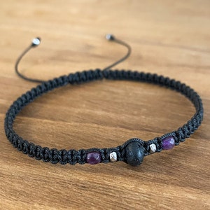 Op de afbeelding: Zwart macramé armband met een centrale zwarte stenen kraal, geflankeerd door twee paarse kralen en zilverkleurige kralen. De armband is verstelbaar met zwarte koorden en wordt gepresenteerd op een houten oppervlak.