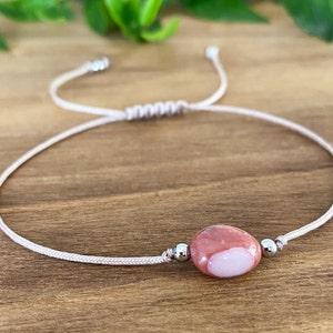 May include: A white string bracelet with a pink and white oval stone bead and silver metal beads.