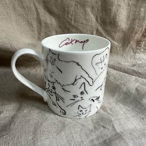Op de afbeelding: Witte keramische mok met een zwarte lijntekening van meerdere katten. De mok heeft een zwart handvat en de tekst "Catnap" in maroonkleurige letters.