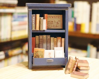 May include: A miniature navy blue bookshelf filled with tiny books of various sizes and colors. The bookshelf has two shelves and a drawer. Some books have visible titles like "FACES" and "AVWXYZ". A small stack of books rests in front of the bookshelf.