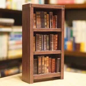 Może przedstawiać: Drewniana, ciemnobrązowa biblioteczka z trzema półkami, każda wypełniona różnymi książkami w skórzanych oprawach. Książki są ułożone w sposób schludny i uporządkowany.